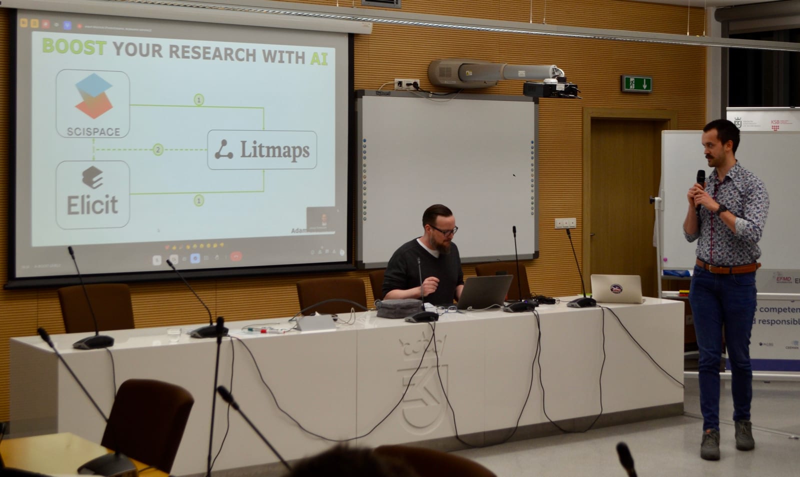 Zdjęcie z sali wykładowej: na ekranie slajd BOOST YOUR RESEARCH WITH AI ze schematem łączącym narzędzia SciSpace, Elicit oraz Litmaps. Przy stole prezydialnym siedzi mężczyzna z laptopem, a z prawej strony stoi drugi z mikrofonem w dłoni.