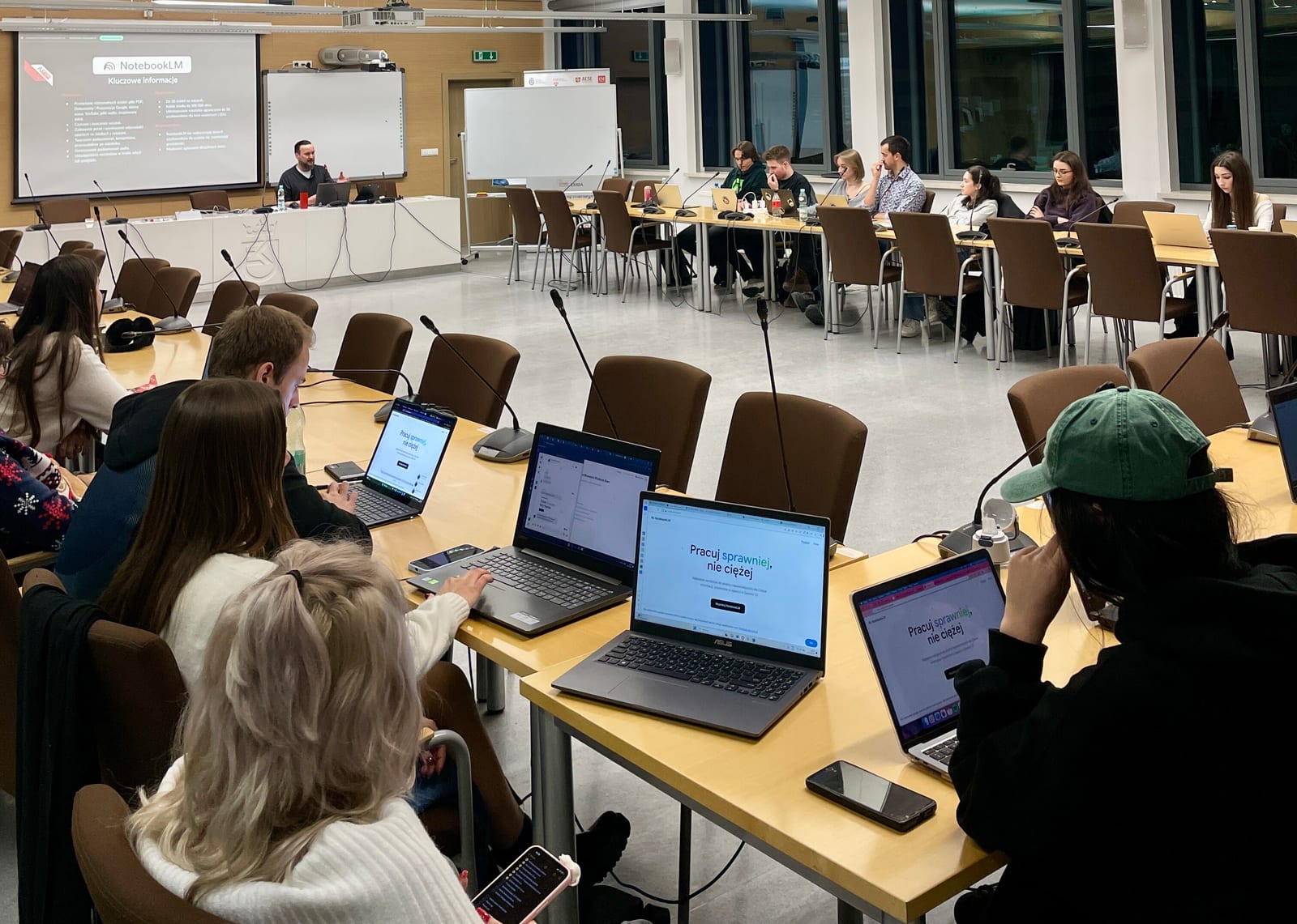 Zdjęcie sali wykładowej od tyłu: uczestnicy siedzą przy stołach z otwartymi laptopami, na których widnieje strona z hasłem Pracuj sprawniej, nie ciężej. Na ekranie w głębi slajd o kluczowych informacjach o NotebookLM, a przy stole prezydialnym prowadzący.
