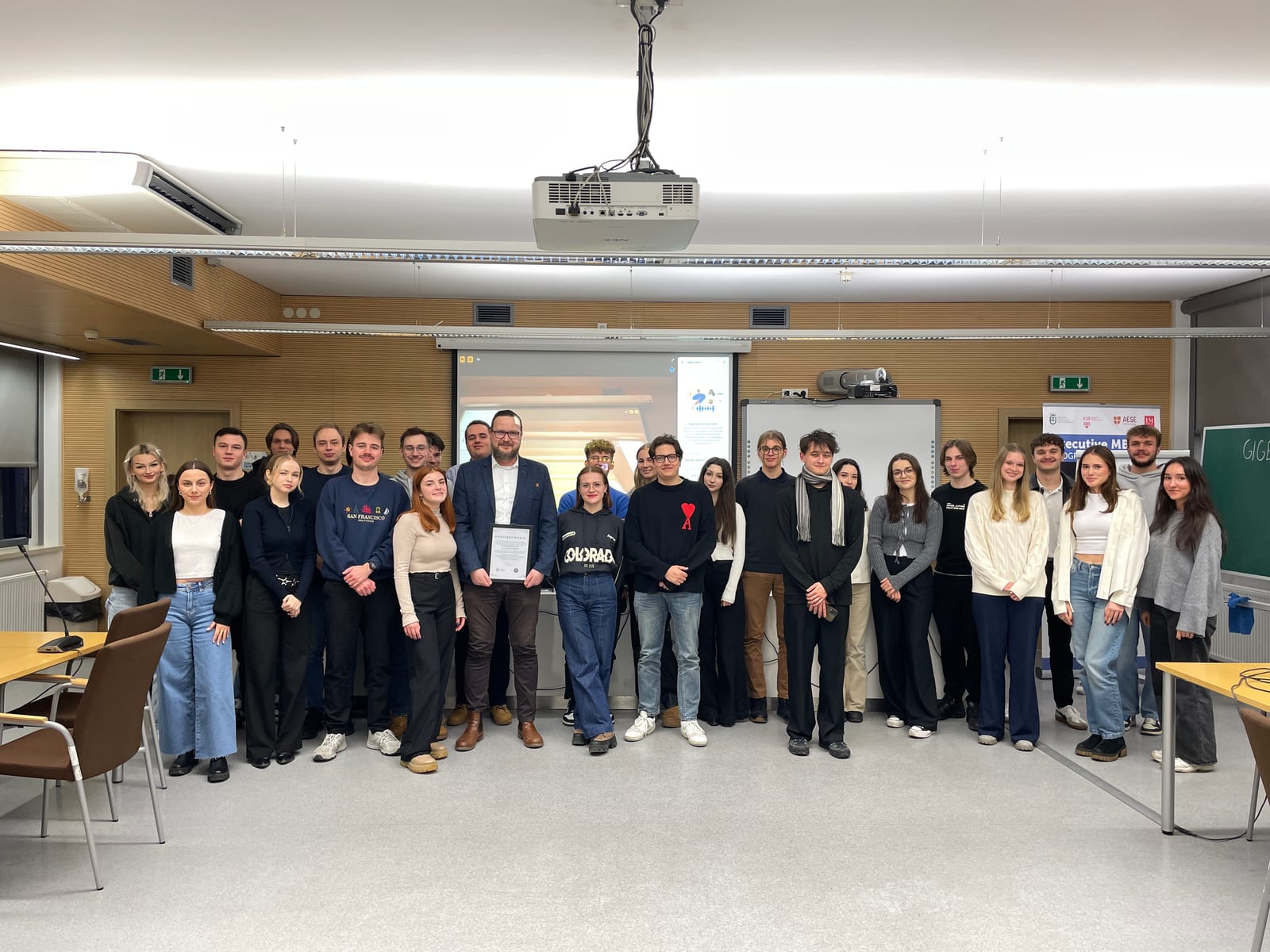 Group photo in a lecture room: about twenty training participants stand in a row, with the presenter in the middle holding a framed letter of thanks. A projection screen, a whiteboard and an Executive MBA banner are visible behind them.