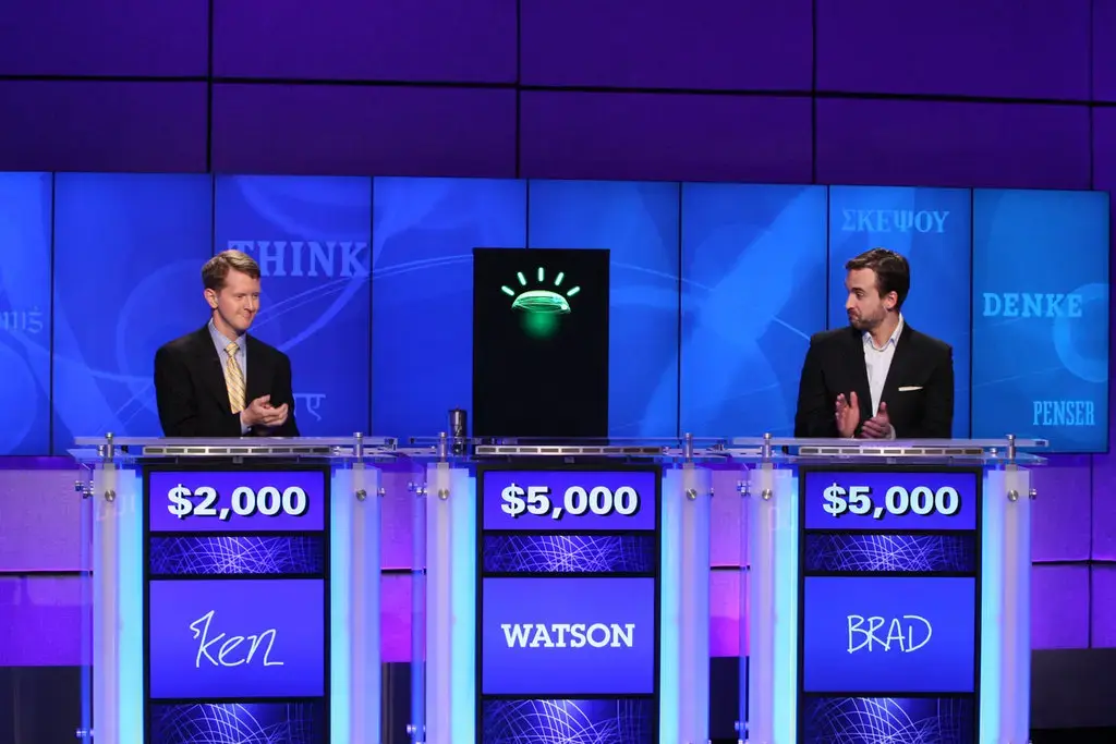 Zdjęcie ze studia teleturnieju Jeopardy: dwaj zawodnicy stoją po bokach, a między nimi pulpit komputera Watson z zielonym znakiem. Na tablicach widnieją wyniki, po 5000 dolarów dla Watsona i Brada oraz 2000 dolarów dla Kena.