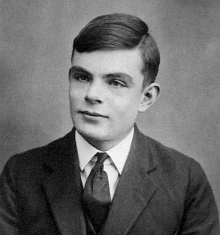 Czarno-biały portret archiwalny: nastoletni Alan Turing w marynarce, koszuli i krawacie, ujęty od pasa w górę na jasnym tle.