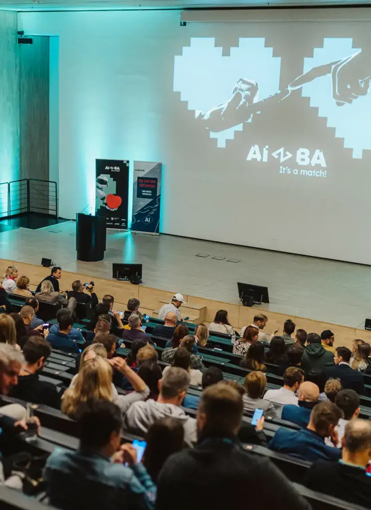 Zdjęcie sali konferencyjnej AIBA widzianej z góry: publiczność wypełnia rzędy krzeseł, część osób fotografuje scenę telefonami, a na dużym ekranie widnieje pikselowe serce, w którym dłoń robota i dłoń ludzka sięgają ku sobie, oraz napis „It’s a match!”.