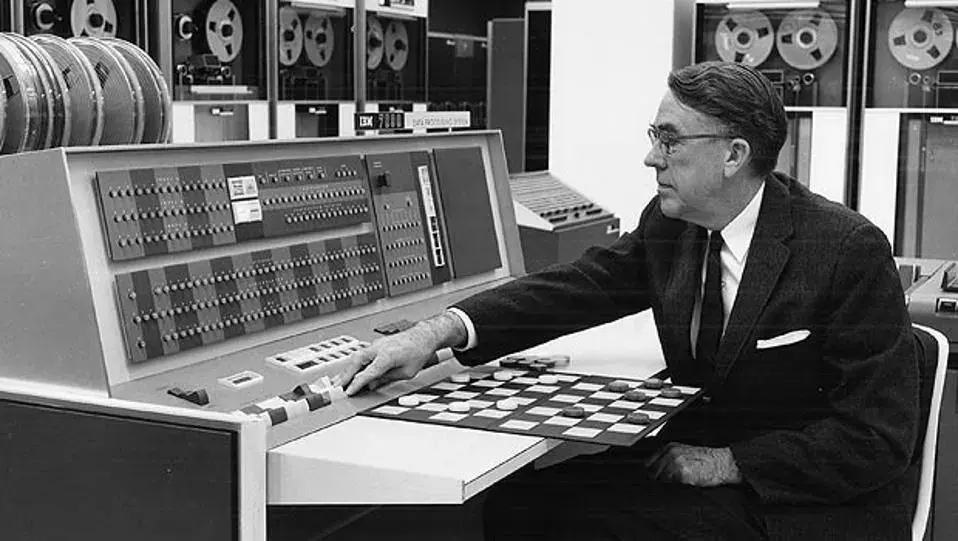 Archival black and white photo: a man in a suit sits at the control desk of a 1960s IBM computer and moves a piece on a checkers board, with tape storage cabinets in the background.