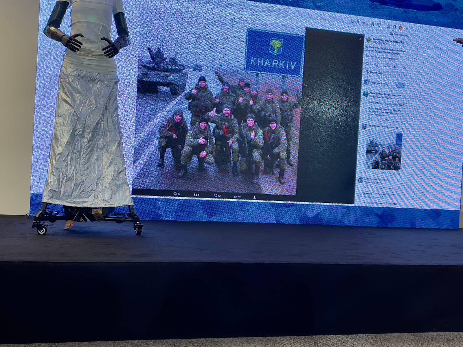 Zdjęcie ze sceny konferencyjnej: ten sam robot stoi przy ekranie, na którym wyświetlono wpis z portalu społecznościowego ze zdjęciem żołnierzy pozujących przy drogowskazie z napisem KHARKIV, z czołgiem w tle.
