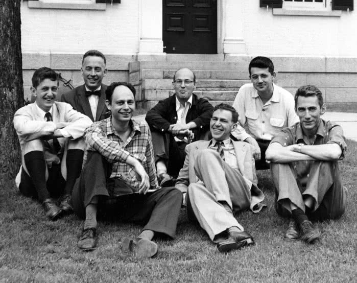 Archival black and white photo of seven men sitting on the grass in front of a stone building, in shirts and jackets, smiling, arranged in two rows. The participants of the 1956 Dartmouth conference.