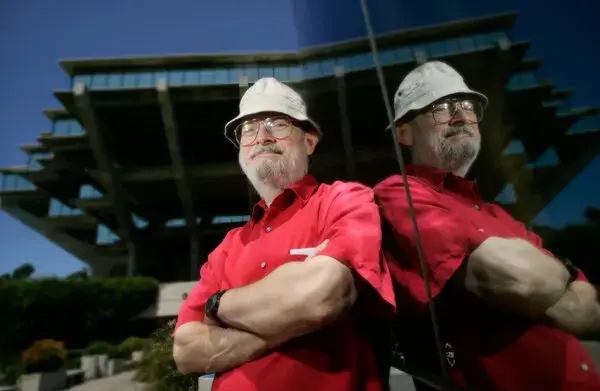 Portrait photo of a bearded man in a red shirt and a white hard hat, arms crossed, against a concrete library building. His figure is reflected in the glass wall on the right of the frame.